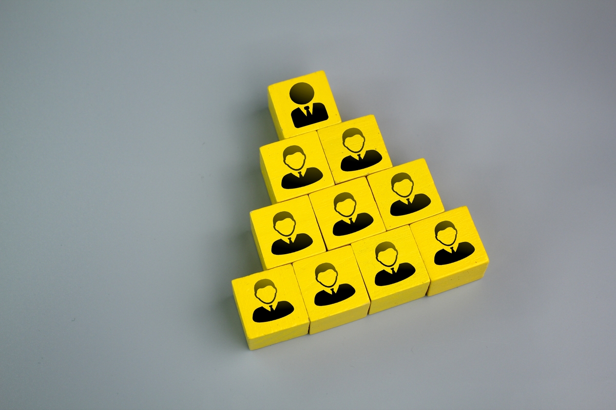 A pyramid of yellow blocks with black silhouettes of professionals, arranged in a hierarchical structure, symbolizes the organizational model of an Intelligent Office franchise. The top block represents a single leader, while the lower rows depict a growing team, set against a plain gray background. Explain hierarchy symbolism Franchise organizational models