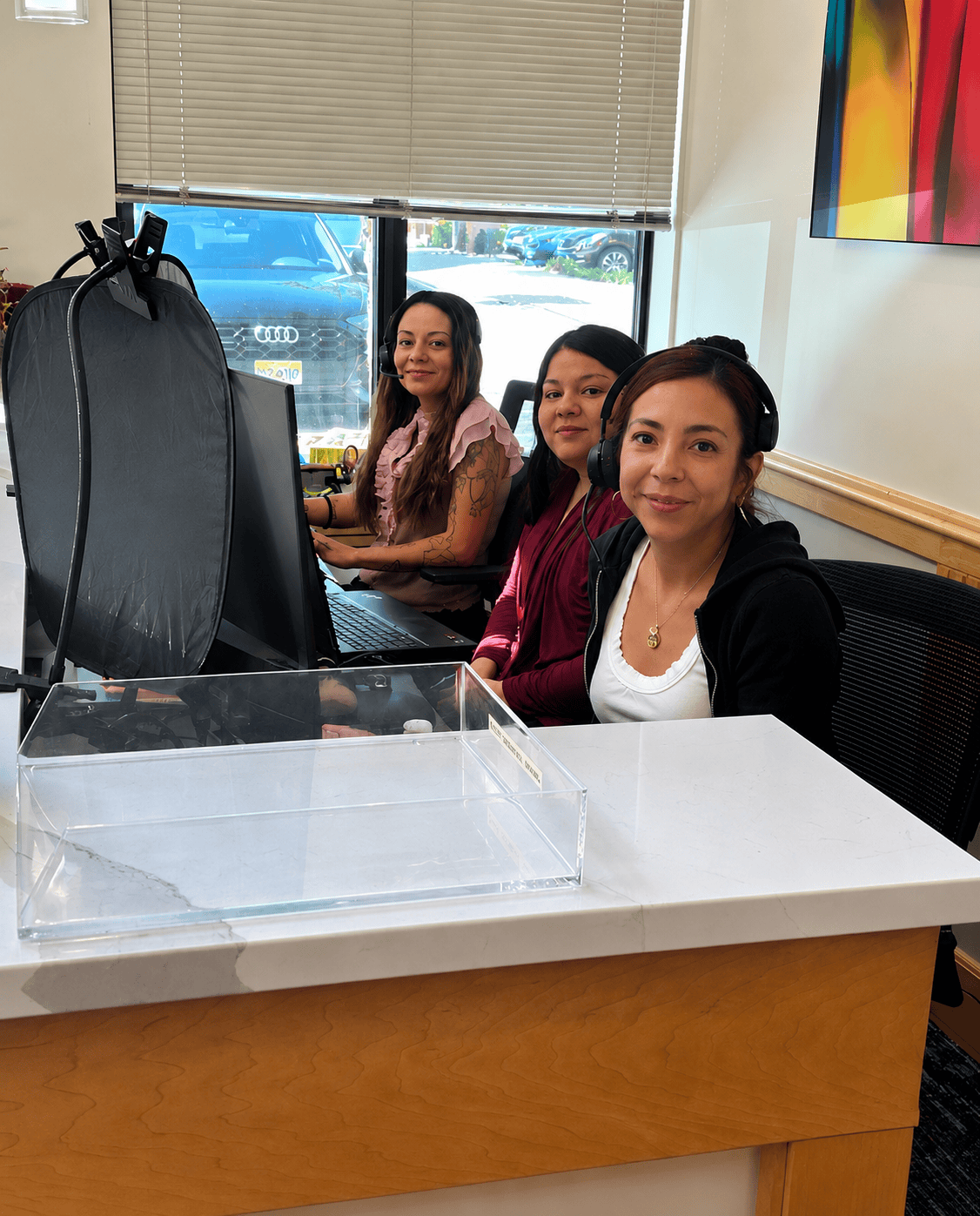 Three women working at a reception desk in an office, smiling and looking at the camera, with a window and cars outside in the background.