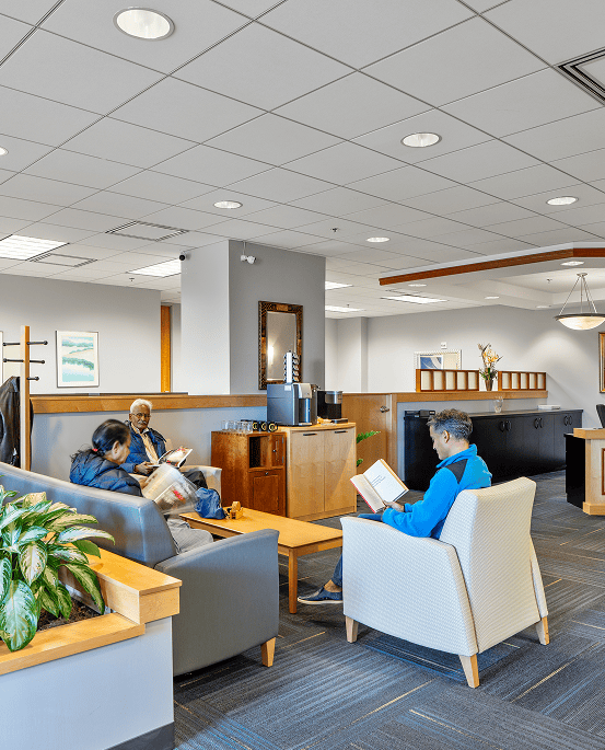 Modern office lounge with clients and staff engaging in discussions and reading, featuring comfortable seating, plants, and contemporary decor in a professional environment.