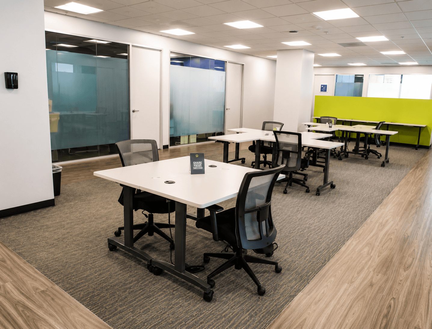 Modern office space with white desks, black chairs, glass walls, and a green accent wall, ideal for collaborative work and meetings.