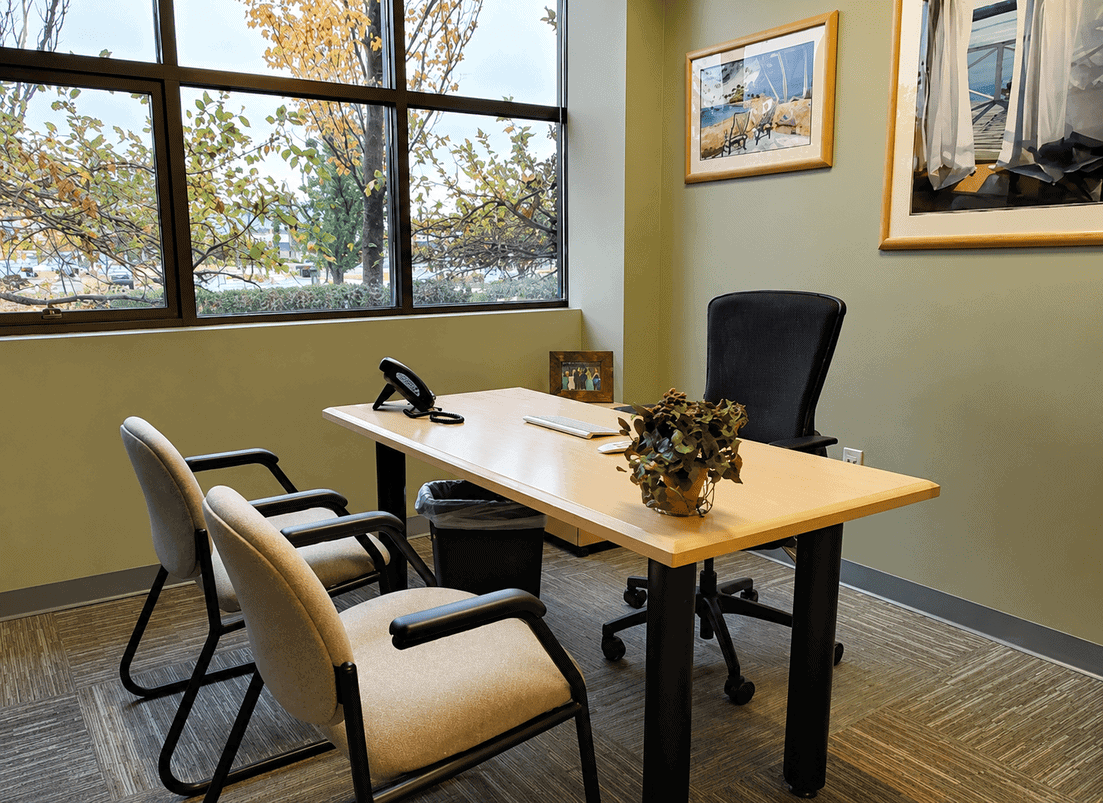 Modern office meeting room with a desk, three chairs, and large windows showing trees outside.
