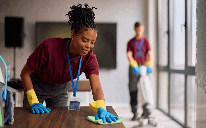 Professional woman cleaning office desk with cloth and gloves, with a janitor in the background, emphasizing office cleaning services.